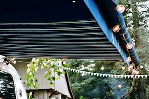 A striped blue awning extends over a scene decorated with string lights. A trailer with a reflective surface is partially visible, alongside some leafy branches and a building. A forest background adds to the outdoor atmosphere.