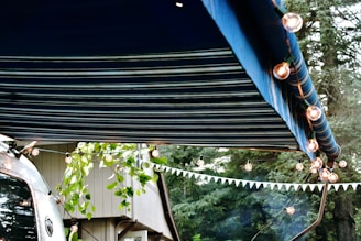 A striped blue awning extends over a scene decorated with string lights. A trailer with a reflective surface is partially visible, alongside some leafy branches and a building. A forest background adds to the outdoor atmosphere.
