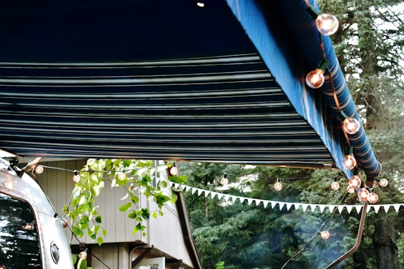 A striped blue awning extends over a scene decorated with string lights. A trailer with a reflective surface is partially visible, alongside some leafy branches and a building. A forest background adds to the outdoor atmosphere.