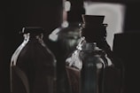 Several glass bottles are arranged closely together, with a dark and moody background. The lighting is soft, casting reflections and shadows on the bottles, highlighting their curves and textures. A cork stopper is visible in the foreground, adding a vintage feel to the scene.