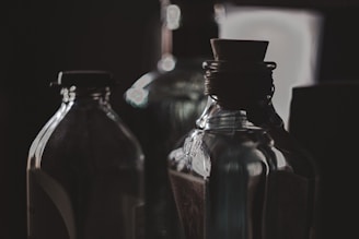 Several glass bottles are arranged closely together, with a dark and moody background. The lighting is soft, casting reflections and shadows on the bottles, highlighting their curves and textures. A cork stopper is visible in the foreground, adding a vintage feel to the scene.