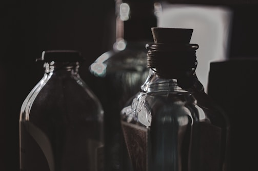 Several glass bottles are arranged closely together, with a dark and moody background. The lighting is soft, casting reflections and shadows on the bottles, highlighting their curves and textures. A cork stopper is visible in the foreground, adding a vintage feel to the scene.