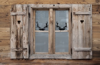 A rustic wooden window with heart-shaped cutouts on each of the two shutters. The window is set in a weathered wooden wall, and through the glass panes, silhouettes of reindeer antlers can be seen, suggesting a decorative motif.