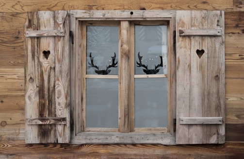A rustic wooden window with heart-shaped cutouts on each of the two shutters. The window is set in a weathered wooden wall, and through the glass panes, silhouettes of reindeer antlers can be seen, suggesting a decorative motif.