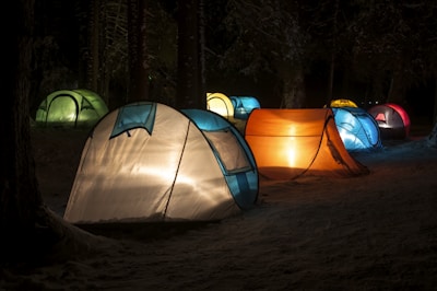 Several brightly lit tents are set up in a dark, wooded area, creating a colorful display against the night backdrop. The tents vary in colors, including shades of blue, orange, green, and red, and they are surrounded by tall, shadowy trees and a dusting of snow on the ground.