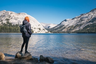 woman wearing black bubble jacket beside lake near hills covered with snow during daytime