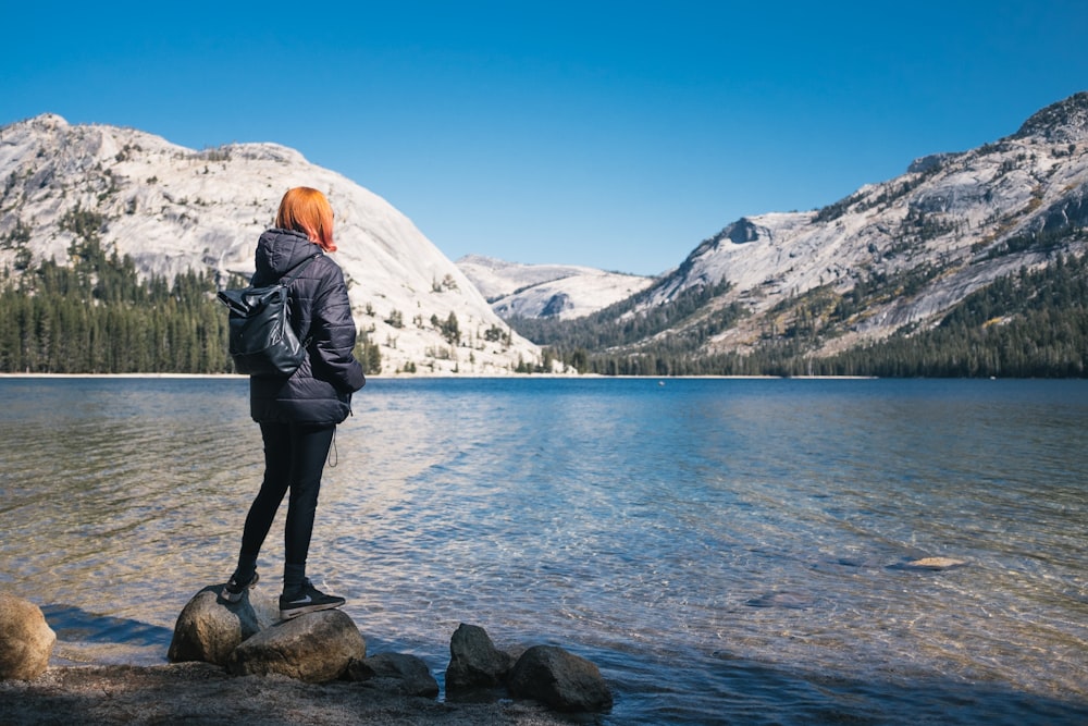 woman wearing black bubble jacket beside lake near hills covered with snow during daytime