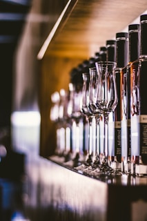 A row of glass bottles displayed on a wooden shelf, accompanied by elegant wine glasses. The setup is well-lit, creating reflections and highlights on the glass surfaces. The background has a soft, blurred effect, focusing attention on the bottles and glasses.
