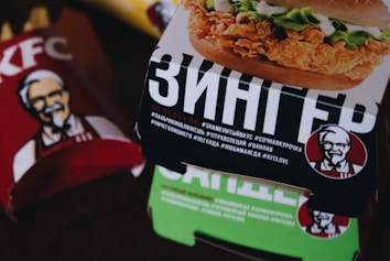 A close-up of KFC packaging, featuring a Zinger burger box prominently in the foreground with Russian text and hashtags. The Colonel Sanders logo is visible on both the Zinger box and the red KFC bag in the background.
