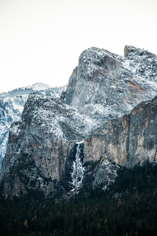 A majestic mountain landscape featuring rocky peaks covered in a light dusting of snow. A narrow waterfall cascades down the cliff face, surrounded by dense coniferous forests at the base.