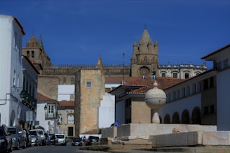 A historic and picturesque street scene features an assortment of white-walled buildings surrounding a central plaza. A prominent old stone cathedral with distinctive towers rises in the background, showcasing Gothic architecture. Parked cars line the street, and a spherical stone fountain stands in the foreground.