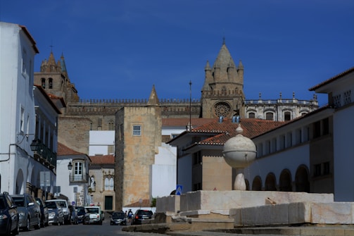 A historic and picturesque street scene features an assortment of white-walled buildings surrounding a central plaza. A prominent old stone cathedral with distinctive towers rises in the background, showcasing Gothic architecture. Parked cars line the street, and a spherical stone fountain stands in the foreground.