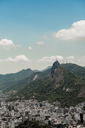 An urban landscape featuring a sprawling city with numerous skyscrapers and high-rise buildings surrounded by lush green hills. The famous Christ the Redeemer statue is seen atop a prominent peak in the background, overlooking the city. The sky is mostly clear with scattered clouds.
