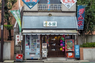 A small shop with a corrugated metal awning, displaying various bright-colored banners with Japanese writing. The exterior includes vending machines, signs, and colorful decorative balls. The building looks aged, with a mix of retro and urban elements surrounded by greenery.