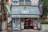 A small shop with a corrugated metal awning, displaying various bright-colored banners with Japanese writing. The exterior includes vending machines, signs, and colorful decorative balls. The building looks aged, with a mix of retro and urban elements surrounded by greenery.