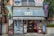 A small shop with a corrugated metal awning, displaying various bright-colored banners with Japanese writing. The exterior includes vending machines, signs, and colorful decorative balls. The building looks aged, with a mix of retro and urban elements surrounded by greenery.