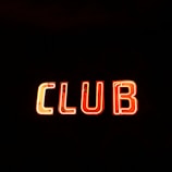 Neon sign displaying the word 'CLUB' in bright, glowing red-orange letters against a stark black background.