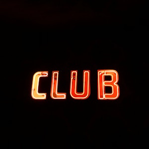 Neon sign displaying the word 'CLUB' in bright, glowing red-orange letters against a stark black background.