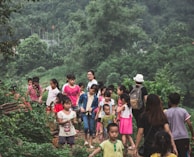 A group of children and a few adults are walking along a path surrounded by lush greenery. The children are wearing casual outfits, with some sporting bright colors. The forested background is dense with a variety of trees and foliage, creating a serene and natural setting.