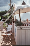 A rustic outdoor setting featuring a canopy-covered bar adorned with greenery and bottles. Director-style chairs with branding are arranged in the foreground, and tents are visible in the background, giving a festival or casual event vibe.