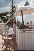 A rustic outdoor setting featuring a canopy-covered bar adorned with greenery and bottles. Director-style chairs with branding are arranged in the foreground, and tents are visible in the background, giving a festival or casual event vibe.