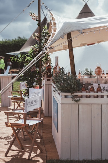 A rustic outdoor setting featuring a canopy-covered bar adorned with greenery and bottles. Director-style chairs with branding are arranged in the foreground, and tents are visible in the background, giving a festival or casual event vibe.