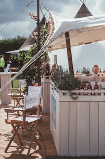 A rustic outdoor setting featuring a canopy-covered bar adorned with greenery and bottles. Director-style chairs with branding are arranged in the foreground, and tents are visible in the background, giving a festival or casual event vibe.