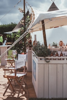 A rustic outdoor setting featuring a canopy-covered bar adorned with greenery and bottles. Director-style chairs with branding are arranged in the foreground, and tents are visible in the background, giving a festival or casual event vibe.