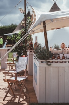 A rustic outdoor setting featuring a canopy-covered bar adorned with greenery and bottles. Director-style chairs with branding are arranged in the foreground, and tents are visible in the background, giving a festival or casual event vibe.