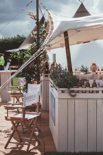 A rustic outdoor setting featuring a canopy-covered bar adorned with greenery and bottles. Director-style chairs with branding are arranged in the foreground, and tents are visible in the background, giving a festival or casual event vibe.