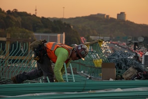 A construction worker wearing a safety vest and helmet is bent over working on a bridge or highway construction site. Green rebar is organized in front of them, with piles of chain link material and cardboard boxes nearby. In the background, a busy highway is visible, with cars lined up in traffic during sunset, casting a warm orange glow over the scene. Hills and modern buildings are visible in the far distance.