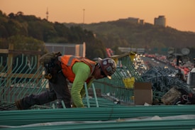 A construction worker wearing a safety vest and helmet is bent over working on a bridge or highway construction site. Green rebar is organized in front of them, with piles of chain link material and cardboard boxes nearby. In the background, a busy highway is visible, with cars lined up in traffic during sunset, casting a warm orange glow over the scene. Hills and modern buildings are visible in the far distance.