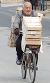 An older man is riding a bicycle loaded with wooden crates on an urban street. He appears to be carrying a heavy load, with crates stacked on the back and front of the bicycle. The street is paved and there are some pedestrians in the background.