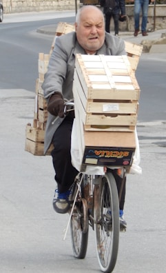 An older man is riding a bicycle loaded with wooden crates on an urban street. He appears to be carrying a heavy load, with crates stacked on the back and front of the bicycle. The street is paved and there are some pedestrians in the background.
