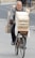 An older man is riding a bicycle loaded with wooden crates on an urban street. He appears to be carrying a heavy load, with crates stacked on the back and front of the bicycle. The street is paved and there are some pedestrians in the background.