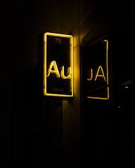 A glowing neon sign against a dark backdrop, displaying the element symbol 'Au' with the atomic number 79 and atomic weight 196.97 in bright yellow. The sign is mirrored on a reflective surface nearby.