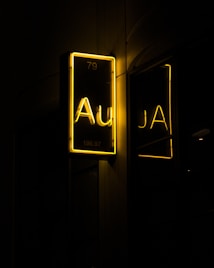 A glowing neon sign against a dark backdrop, displaying the element symbol 'Au' with the atomic number 79 and atomic weight 196.97 in bright yellow. The sign is mirrored on a reflective surface nearby.