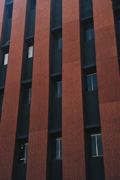 A multi-story building facade with alternating vertical sections of red brick and dark windows. The bricks are laid in a consistent pattern, and the structure has a modern, geometric appearance.