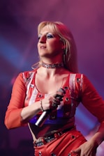 A person with long blonde hair is performing on stage, holding a microphone. They are wearing a vibrant red outfit adorned with silver sequins, under dramatic stage lighting that casts purple and blue hues.