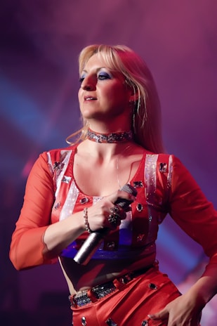 A person with long blonde hair is performing on stage, holding a microphone. They are wearing a vibrant red outfit adorned with silver sequins, under dramatic stage lighting that casts purple and blue hues.