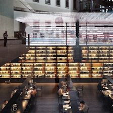 A modern library interior with large bookshelves filled with books. Several people are seated at long tables working on laptops and reading. The space is illuminated by warm lighting, creating a cozy atmosphere. Architectural elements and reflections add a layered effect to the image.