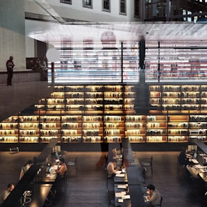 A modern library interior with large bookshelves filled with books. Several people are seated at long tables working on laptops and reading. The space is illuminated by warm lighting, creating a cozy atmosphere. Architectural elements and reflections add a layered effect to the image.