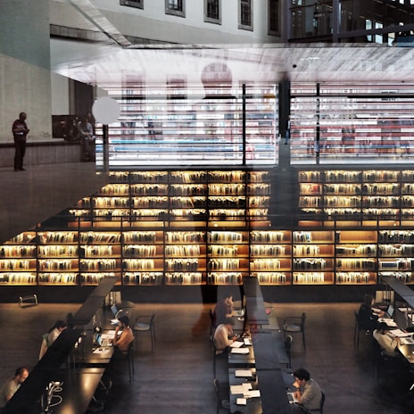 A modern library interior with large bookshelves filled with books. Several people are seated at long tables working on laptops and reading. The space is illuminated by warm lighting, creating a cozy atmosphere. Architectural elements and reflections add a layered effect to the image.
