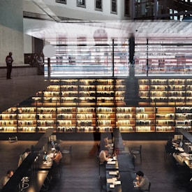 A modern library interior with large bookshelves filled with books. Several people are seated at long tables working on laptops and reading. The space is illuminated by warm lighting, creating a cozy atmosphere. Architectural elements and reflections add a layered effect to the image.