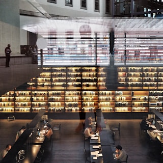 A modern library interior with large bookshelves filled with books. Several people are seated at long tables working on laptops and reading. The space is illuminated by warm lighting, creating a cozy atmosphere. Architectural elements and reflections add a layered effect to the image.