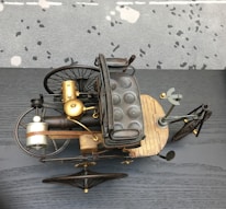 An antique-style tricycle with a wooden base, padded seat, and metal framework is showcased from above. The design includes large, spoked wheels, a front gear mechanism, and brass components, suggesting historical or steampunk aesthetics. The background is a speckled carpet and a dark gray, textured surface.
