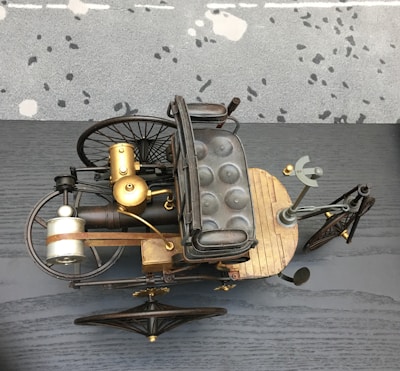 An antique-style tricycle with a wooden base, padded seat, and metal framework is showcased from above. The design includes large, spoked wheels, a front gear mechanism, and brass components, suggesting historical or steampunk aesthetics. The background is a speckled carpet and a dark gray, textured surface.