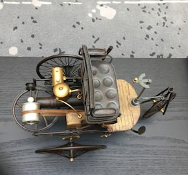 An antique-style tricycle with a wooden base, padded seat, and metal framework is showcased from above. The design includes large, spoked wheels, a front gear mechanism, and brass components, suggesting historical or steampunk aesthetics. The background is a speckled carpet and a dark gray, textured surface.