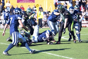 A group of American football players wearing helmets and uniforms are in the middle of a play on a grassy field. One player in a blue and white uniform is on the ground holding the ball, while others in green and black uniforms attempt to tackle him. The scene is dynamic, capturing the action of the game with a focus on the struggle for possession. Spectators are visible in the background.