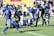 A group of American football players wearing helmets and uniforms are in the middle of a play on a grassy field. One player in a blue and white uniform is on the ground holding the ball, while others in green and black uniforms attempt to tackle him. The scene is dynamic, capturing the action of the game with a focus on the struggle for possession. Spectators are visible in the background.